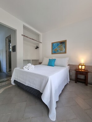 A neatly made bed with white linens and a blue pillow, a small bedside table with a lamp, and a painting on the wall. - Pousada Solar das Orquídeas Praia (Porto Seguro)