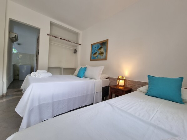 A hotel room with two beds, a small bedside table with a lamp, a painting on the wall, and a bathroom visible through an open door. - Pousada Solar das Orquídeas Praia (Porto Seguro)