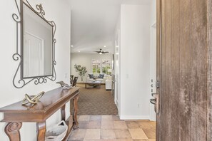Un pasillo con una consola de madera, un espejo decorativo y una puerta que da a una sala de estar con sofá y ventilador de techo.