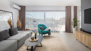 A modern living room with a grey sofa, teal armchair, and a coffee table with a bowl of fruit. There is a large window with sheer curtains, a floor lamp, and a TV mounted on the wall. - Luxury Villa Silvery with Pool (Kanica)