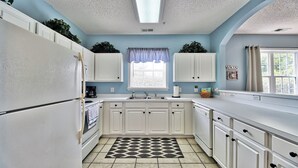 Una cocina con armarios blancos, un tapete con estampado blanco y negro, una ventana con cortina azul y una lámpara en el techo.