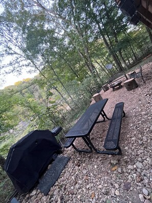 Un espace extérieur aménagé avec des tables de pique-nique et un barbecue, entouré d’arbres et d’un sol rocheux.