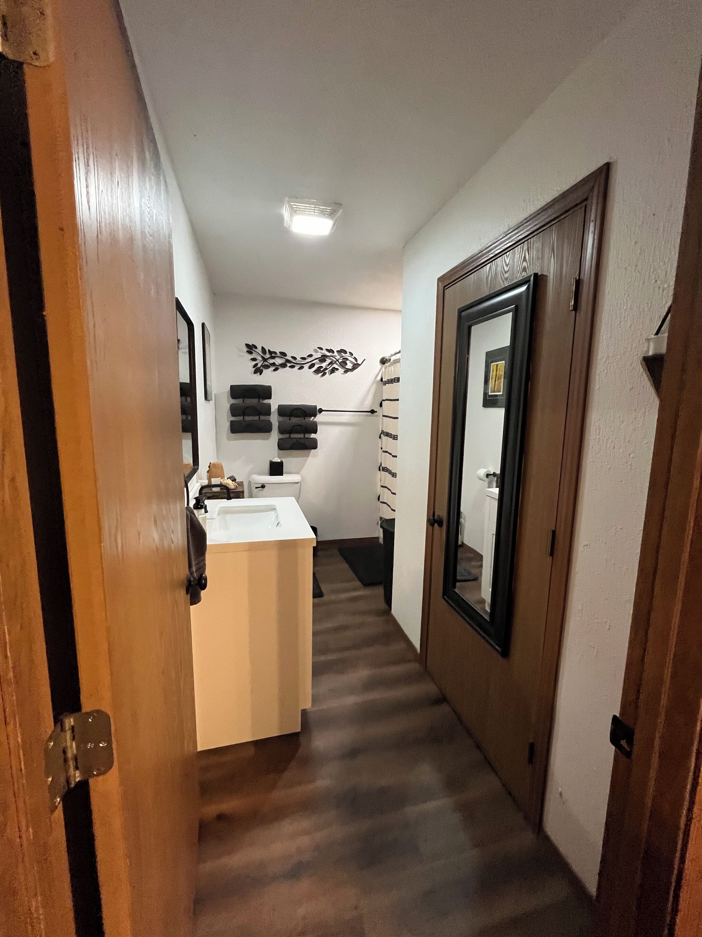 Un couloir étroit avec une porte en bois sur la droite, un lavabo blanc avec un miroir et des toilettes avec un porte-serviettes.