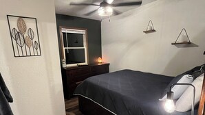 Ein Schlafzimmer mit Bett, Deckenventilator, Nachttisch mit Lampe, Wandregal und Fenster mit Jalousien.