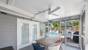 A covered patio area with a table and chairs, a ceiling fan, and a view of a backyard with a pool and lounge chairs. - Luxury Beach House | Private Pool & Walk Everywhere (St. Pete Beach)