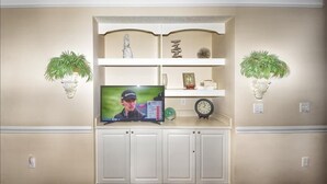 A living room with a TV on a white cabinet, two wall-mounted plants, a clock, and a shelf with decorative items. - 512 The Havens - Barefoot Resort Rentals (North Myrtle Beach)