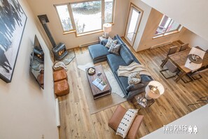 A spacious living room with a blue sofa, wooden flooring, a fireplace, and large windows offering views of the outdoors. - Adorable Townhome! 15 Min Drive to Resort! 5 Min Walk to Town Center and Shuttle (Big Sky)