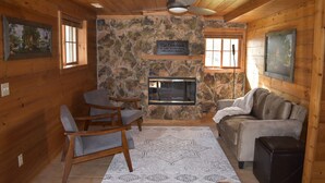 Un intérieur de chalet chaleureux avec une cheminée en pierre, des meubles en bois et une pancarte indiquant : « L’aventure vous attend au point de vue sur le lac Kosihonong ».