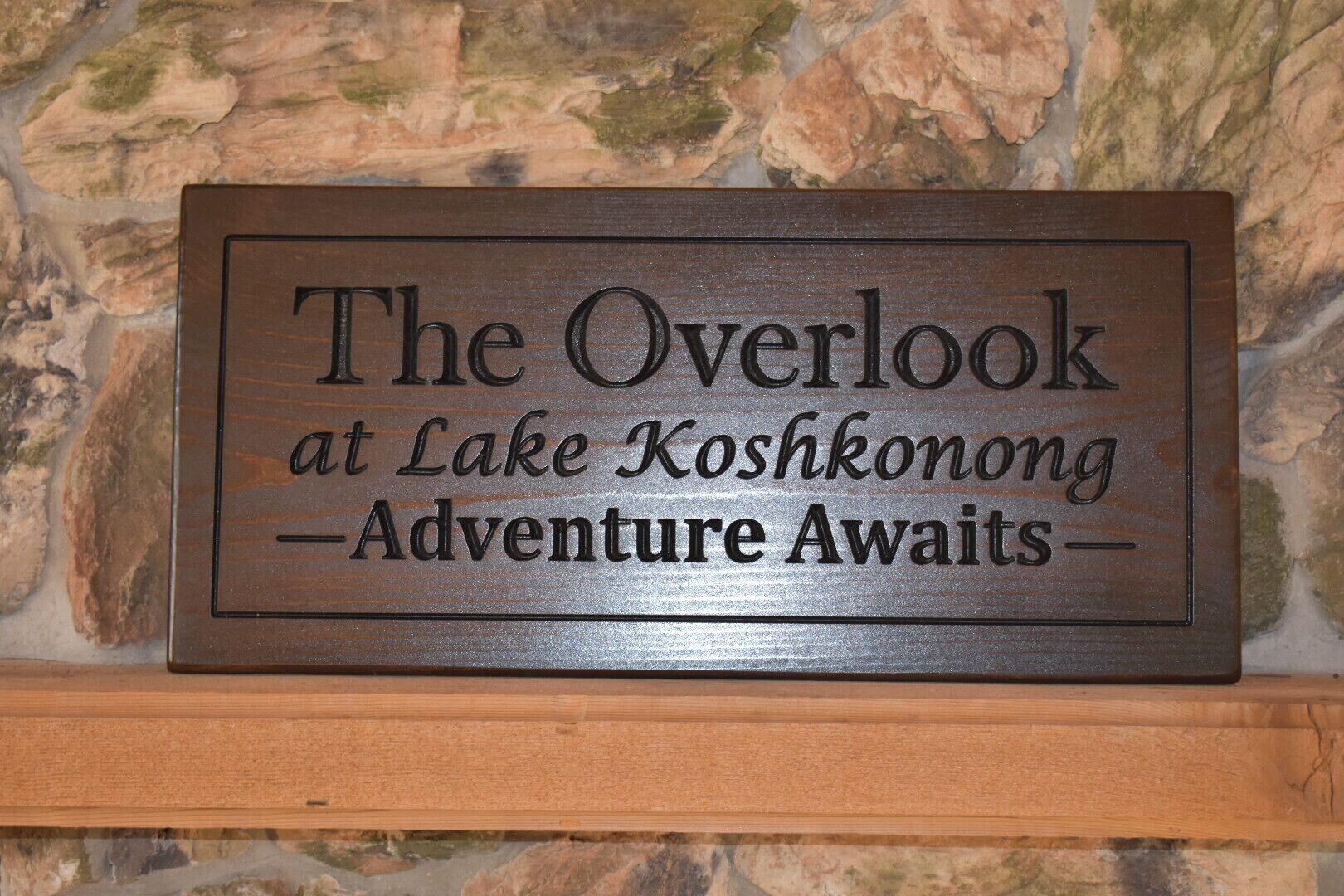 A wooden sign with the text 'The Overlook at Lake Koshkonong - Adventure Awaits' mounted on a stone wall.
