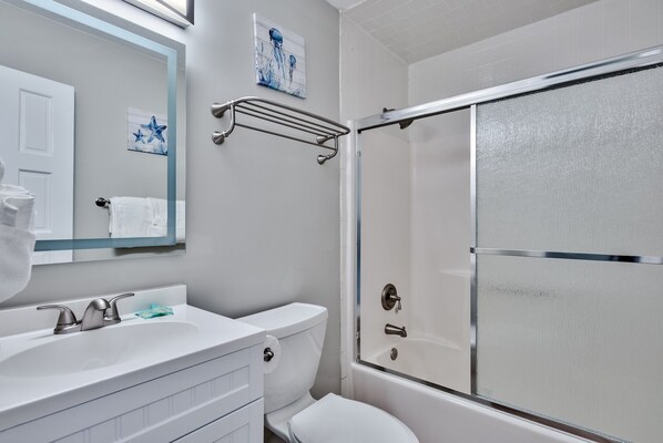 Een badkamer met een wit wastafelmeubel, toilet en douche. Boven het wastafelmeubel hangt een handdoekrek en aan de muur een schilderij.