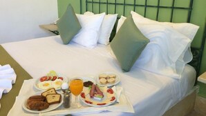 Cama bem arrumada com roupa de cama branca e almofadas verdes, bandeja com café da manhã incluindo ovos, torradas e suco, e parede verde com padrão quadriculado.
