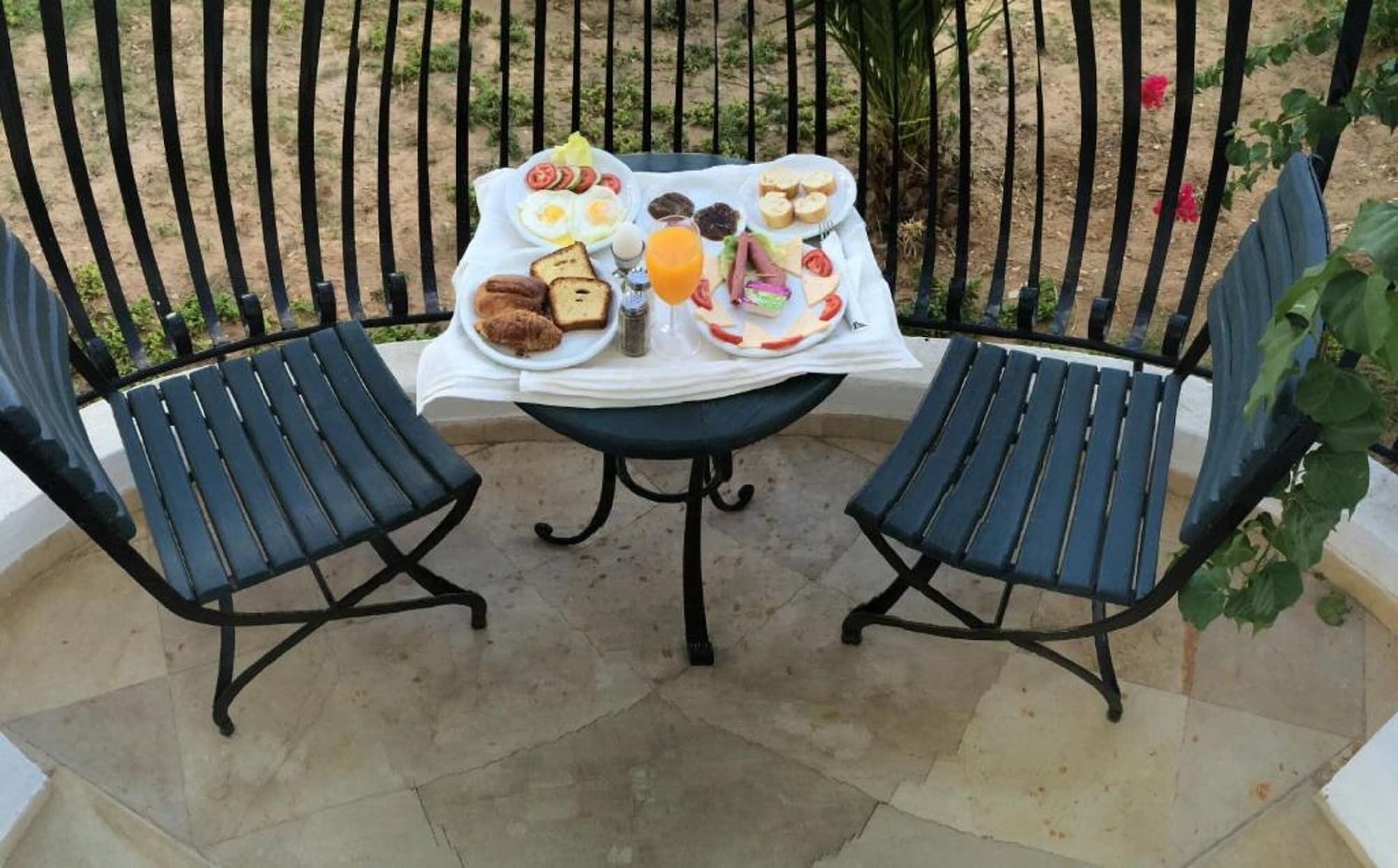 Une table dressée pour deux personnes, avec une variété d’aliments pour le petit-déjeuner, notamment des œufs, des toasts et des fruits, sur une terrasse dotée de chaises et d’une rambarde.