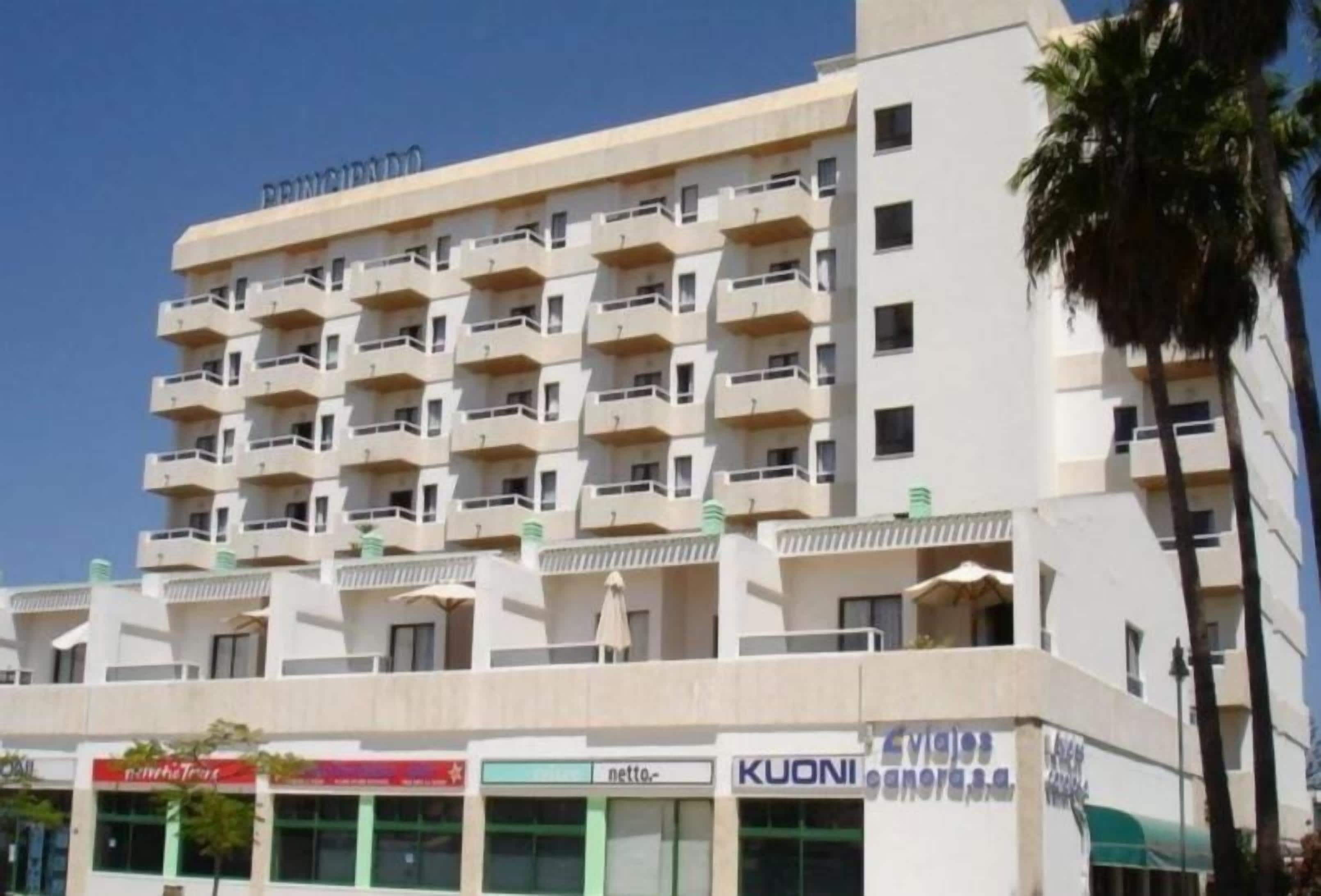 A multi-story hotel with balconies, a ground-level retail area, and palm trees.