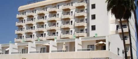A multi-story hotel with balconies, a ground-level retail area, and palm trees.