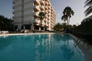 Una piscina con sillones de descanso y un edificio de hotel de varios pisos al fondo.