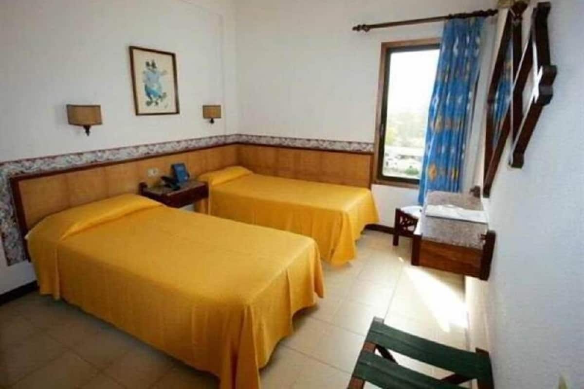 Una habitación con dos camas, una mesita, una silla, un ventanal con cortinas y una lámpara fijada a la pared.