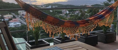 A hammock on a balcony overlooking a cityscape at sunset.