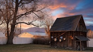 A rustic wooden barn with a sloped roof, surrounded by a fenced yard, and a large tree to the left. - Restored 1920s Corner Cottage – Bryce Gateway Retreat (Panguitch)