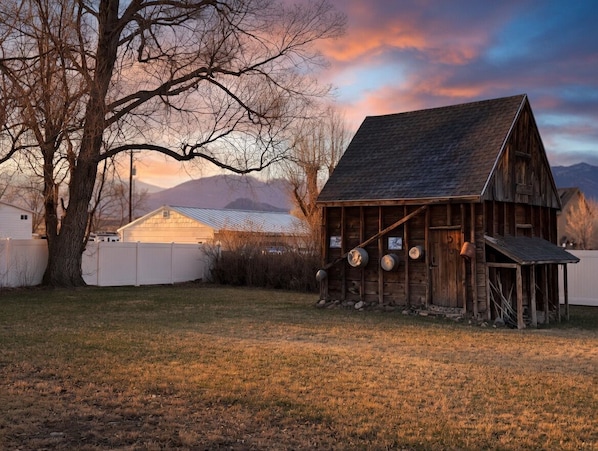 A rustic wooden barn with a sloped roof, surrounded by a fenced yard, and a large tree to the left.