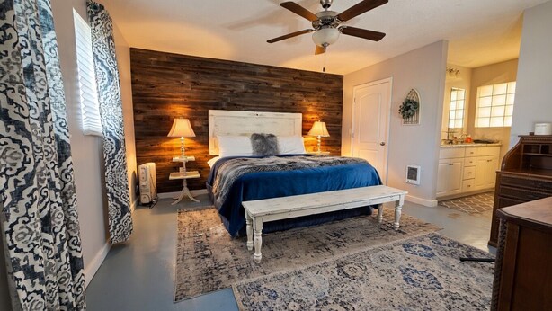 A bedroom with a wooden headboard, a bed with a blue bedspread, a white bench, a ceiling fan, and a view of the kitchen. - Restored 1920s Corner Cottage – Bryce Gateway Retreat (Panguitch)