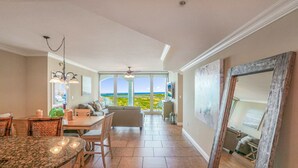 A spacious living area with a large window offering a view of the ocean, a dining area with a table and chairs, and a cozy seating area with a couch and chairs.
