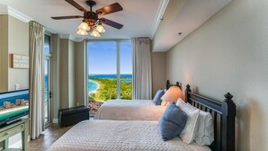 A hotel room with two beds, a ceiling fan, a TV, and a view of the beach.