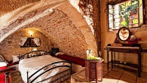 A rustic bedroom with a stone ceiling, a bed with a red blanket, a bedside table with a mirror, and a window with a view of greenery. - Veneto Boutique Hotel (Rethymno)