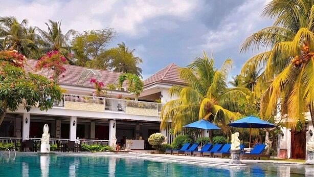 A clear swimming pool with blue lounge chairs and umbrellas, surrounded by palm trees and a building with a balcony. - Puri Saron Senggigi Beach Hotel (Senggigi)