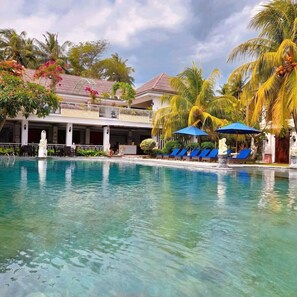 A clear swimming pool with blue lounge chairs and umbrellas, surrounded by palm trees and a building with a balcony. - Puri Saron Senggigi Beach Hotel (Senggigi)