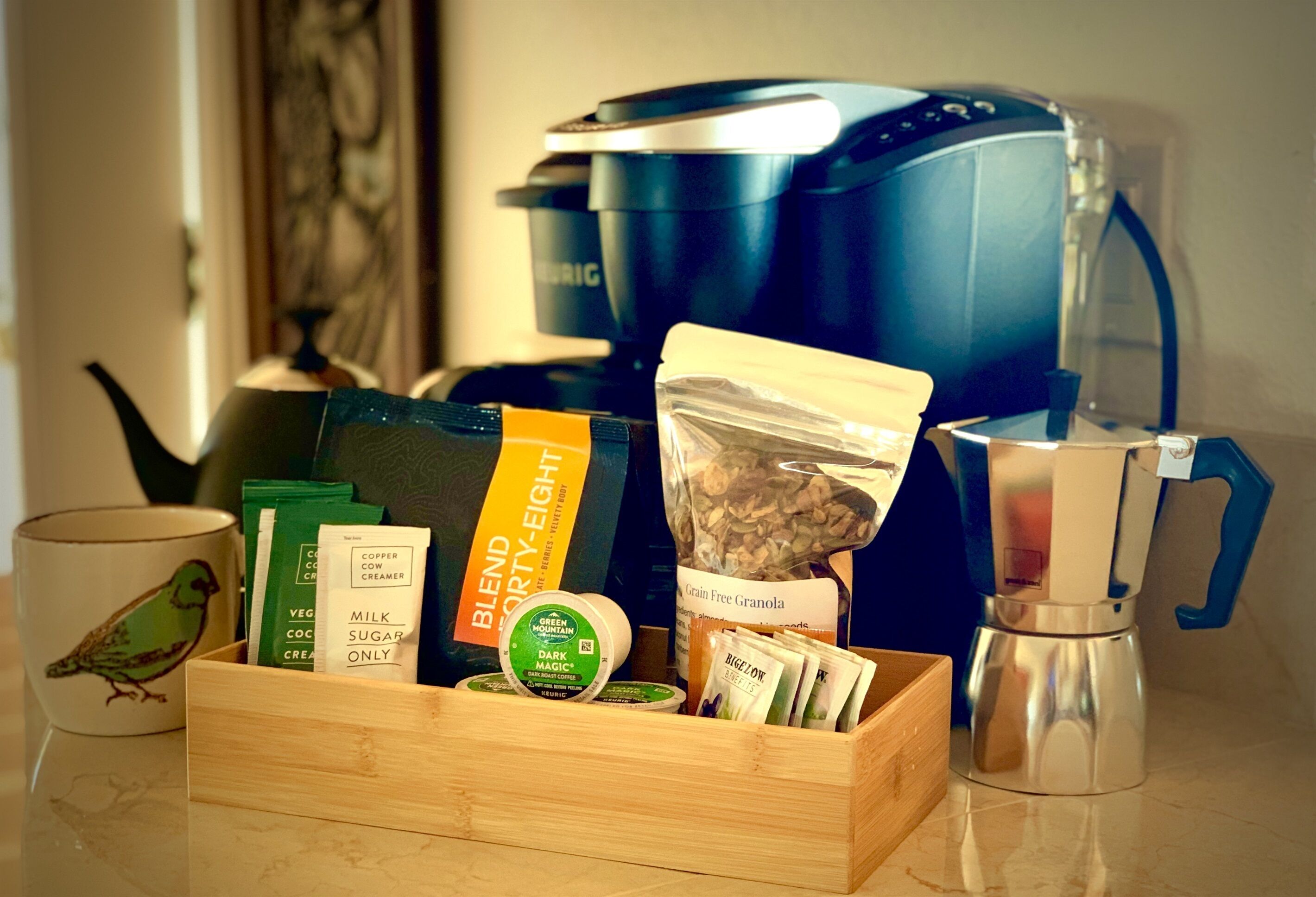 A kitchen counter with a coffee maker, a wooden box containing coffee pods, a teapot, and a cup with a bird design.