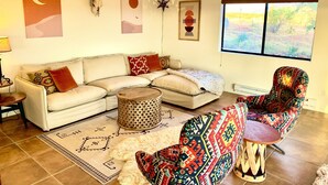 Una sala de estar con un sofá modular, un sillón estampado, una mesa de centro redonda de madera y un tapiz decorativo.