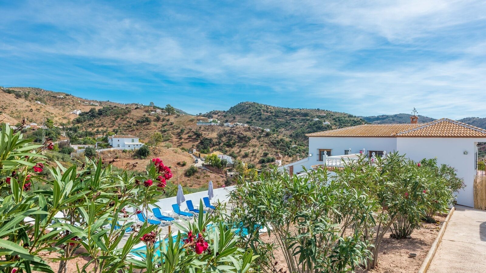Un espace piscine avec des chaises longues bleues, un bâtiment blanc doté d’un toit en tuiles et un paysage vallonné en arrière-plan.