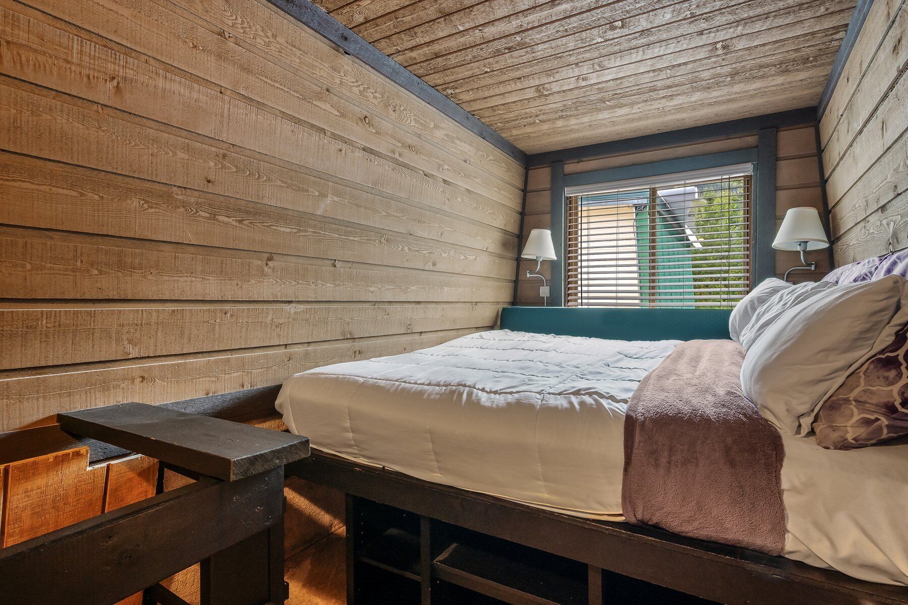 A bedroom with a wooden sloped ceiling, a bed with a white comforter and a brown throw blanket, two bedside lamps, and a window with blinds.