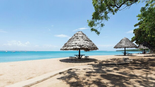 A beach with thatched umbrellas and tables, a clear sky, and the ocean in the background. - Four Points By Sheraton Dar Es Salaam New Africa (Dar es Salaam)