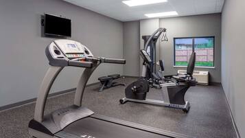 Un gimnasio con cinta de correr, elíptica y máquina de pesas, un televisor en la pared y una ventana con vistas a una valla.