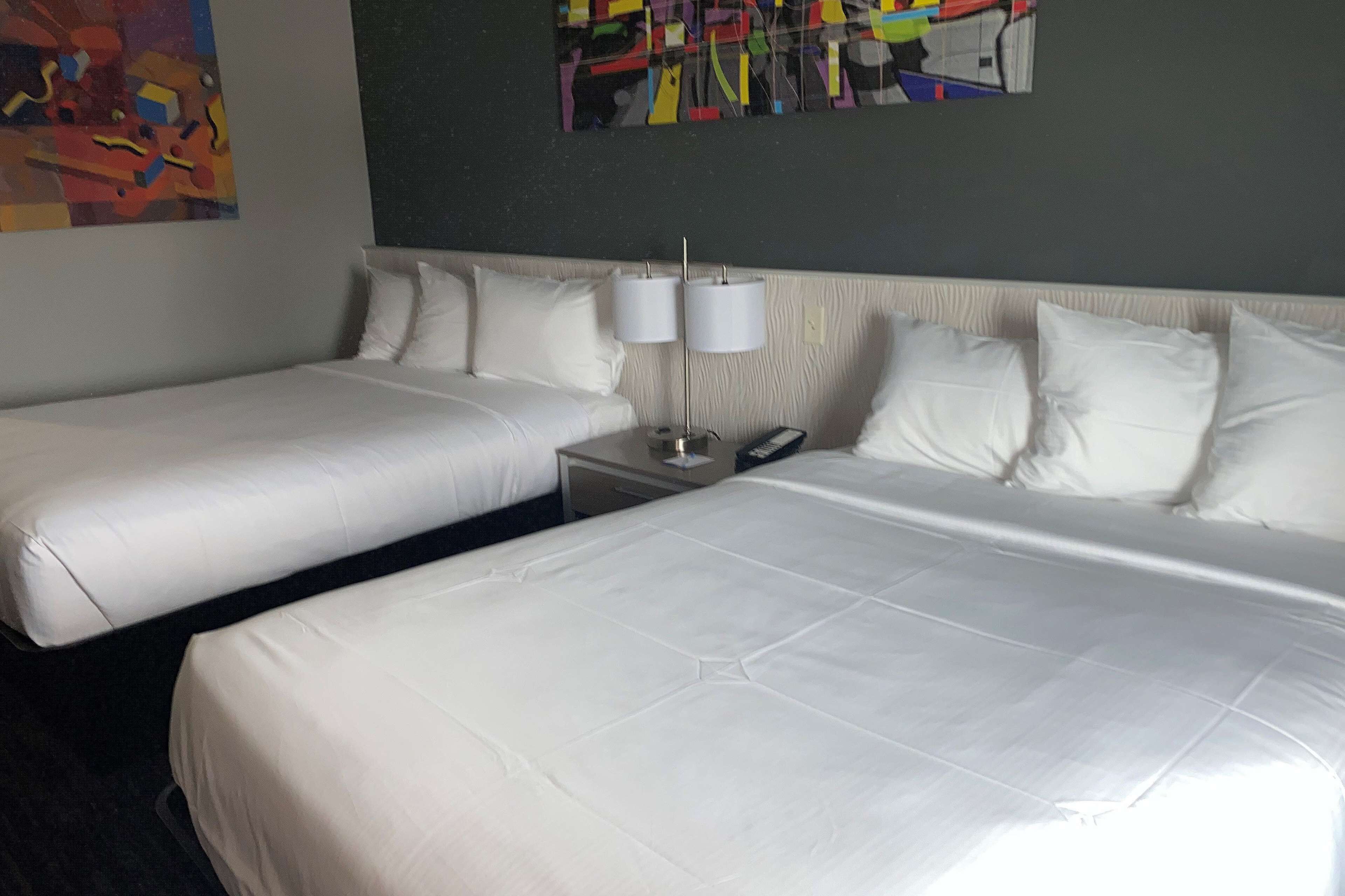 Quarto de hotel com duas camas, roupa de cama branca, mesa de cabeceira com abajur e arte abstrata na parede.