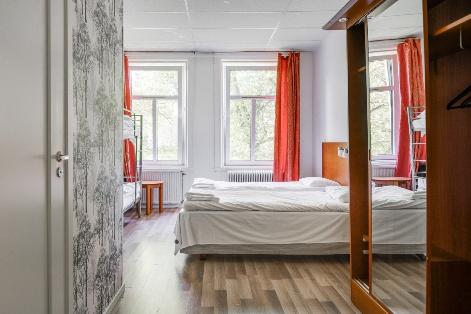 A bedroom with two beds, a wooden floor, a radiator, and a window with red curtains.