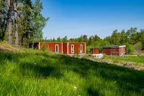 A rustic cabin with a red exterior and white trim, situated in a grassy field with trees in the background.