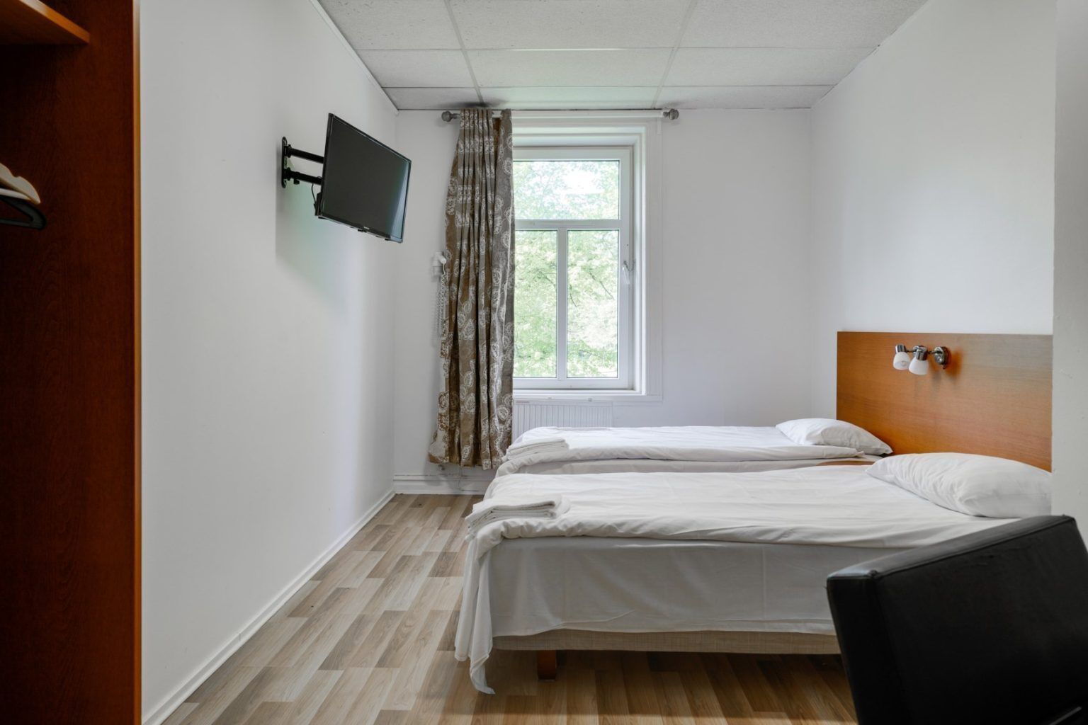 A hotel room with two beds, a TV mounted on the wall, a window with curtains, and a wooden headboard.