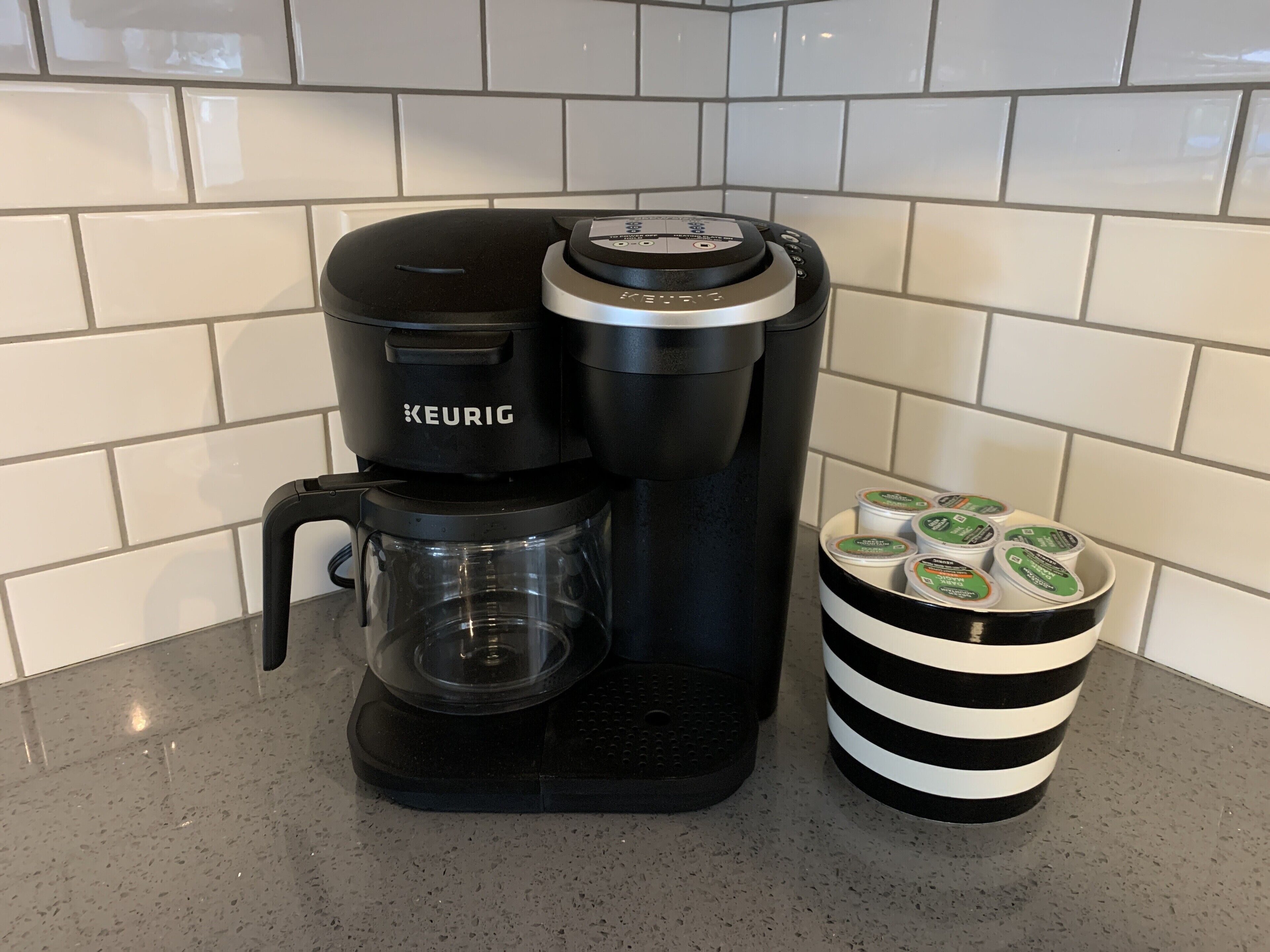 Une machine à café Keurig dotée d’une verseuse en verre et d’une tasse à rayures noires et blanches, remplie de capsules Keurig.