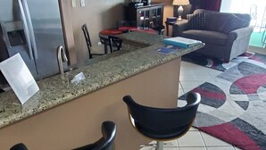 Un área de cocina con refrigerador, microondas y cafetera. Hay una mesa de comedor con sillas y una barra con taburetes. Un televisor está montado en la pared sobre la barra.