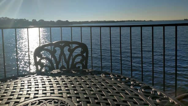 Una mesa metálica con un diseño trenzado, una baranda de metal y vista a un cuerpo de agua con una orilla lejana.