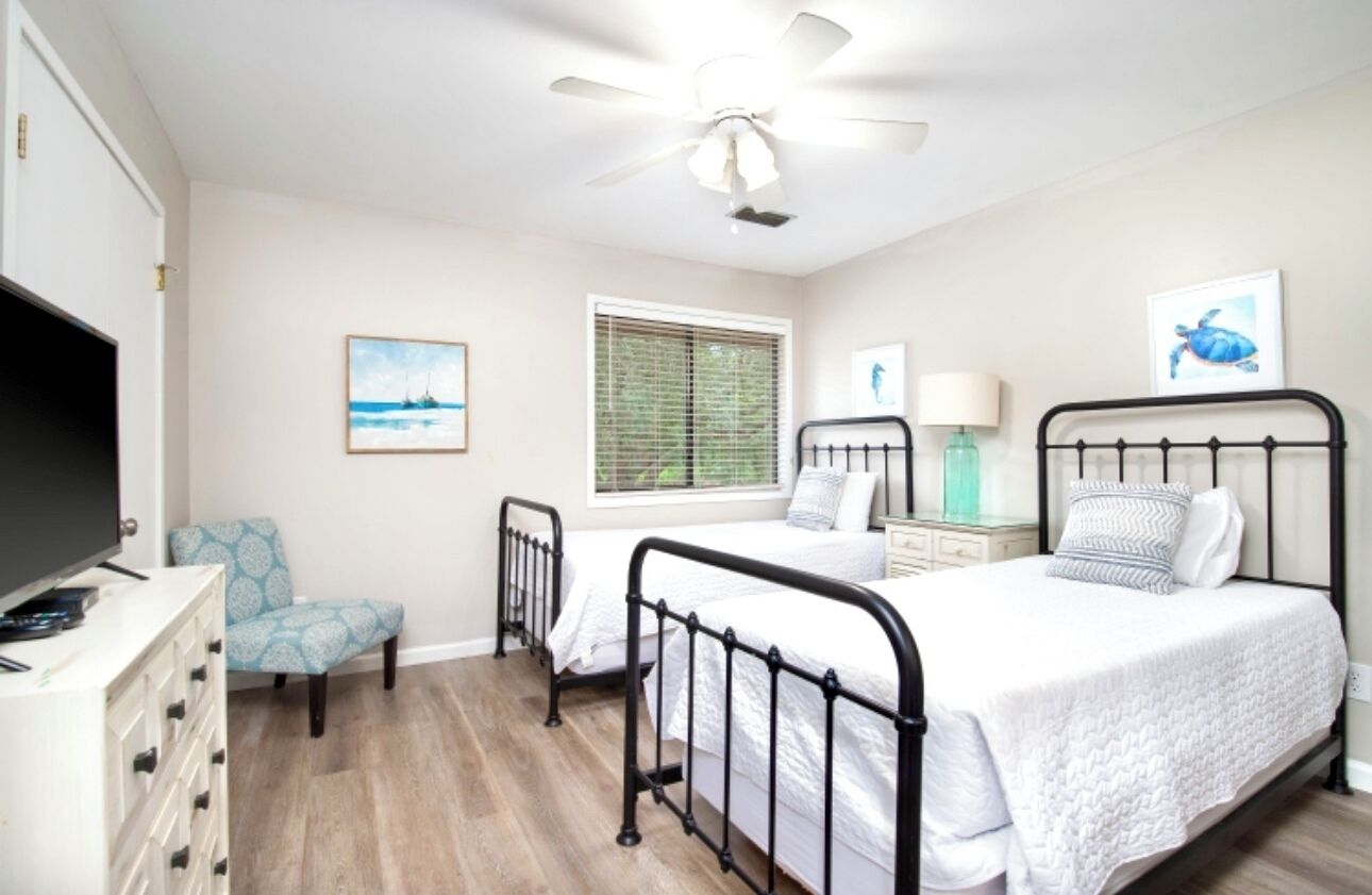 A bedroom with two beds, a ceiling fan, a TV, a chair, and artwork on the walls.