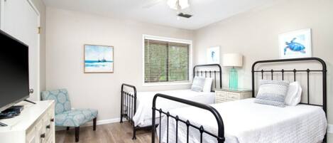 A bedroom with two beds, a ceiling fan, a TV, a chair, and artwork on the walls.