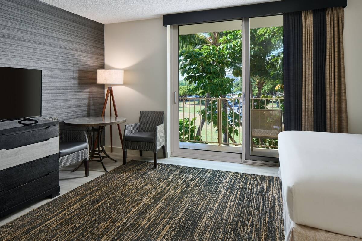 Habitación de hotel con televisión, una mesa de comedor con sillas, un sofá y un ventanal con vistas a palmeras y un estacionamiento.