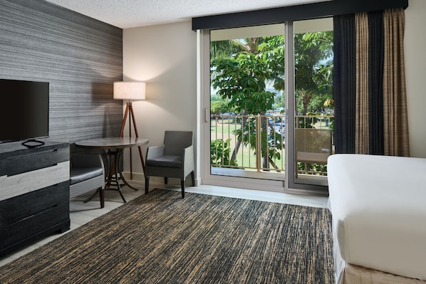 Habitación de hotel con televisión, una mesa de comedor con sillas, un sofá y un ventanal con vistas a palmeras y un estacionamiento.