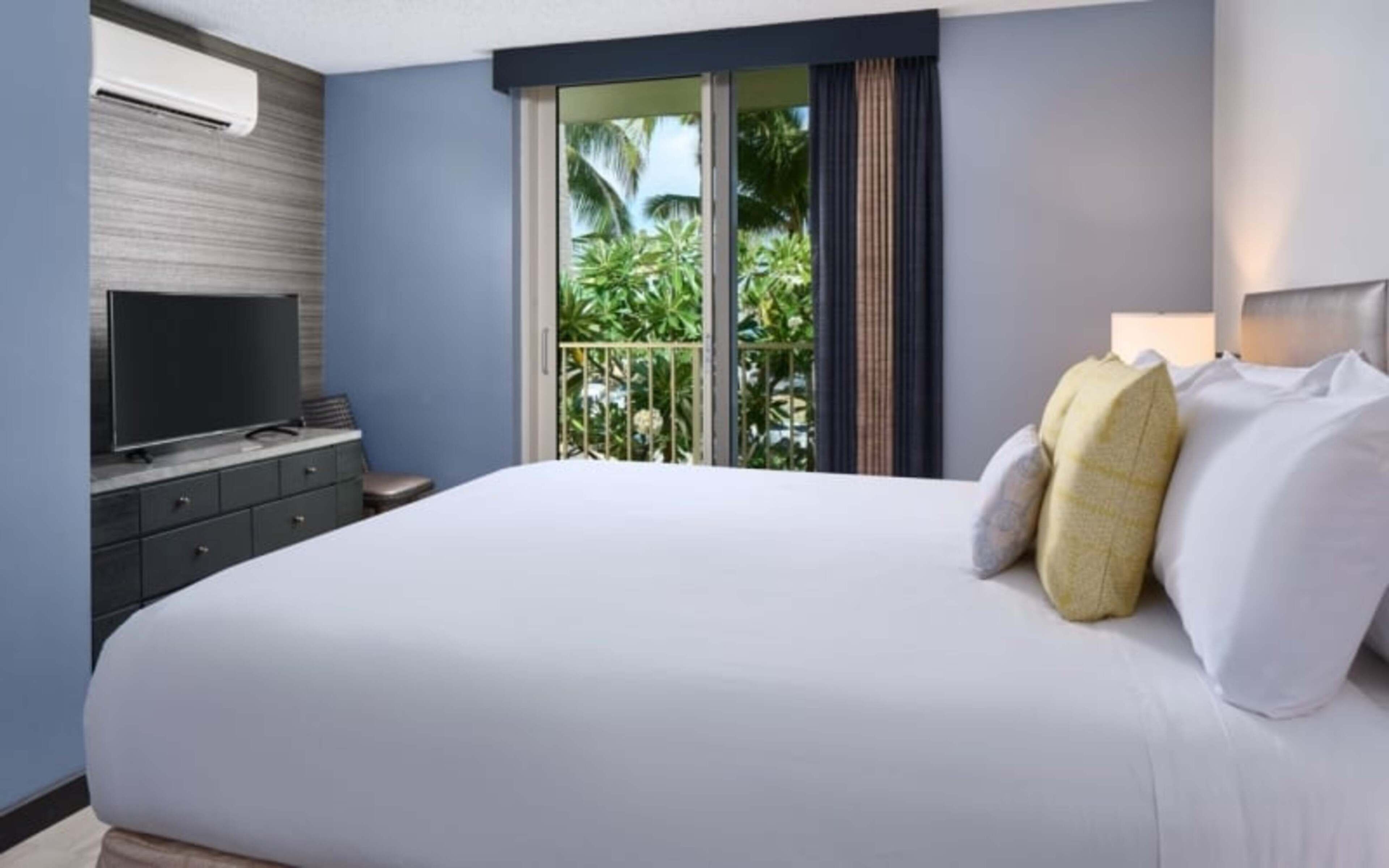 Una habitación de hotel con una cama grande, un televisor de pantalla plana sobre un aparador y vistas a palmeras a través de la ventana.