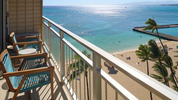 A balcony with wooden chairs and a table overlooking a beach with clear turquoise water and a sandy shore. - Kaimana Beach Hotel (Honolulu)