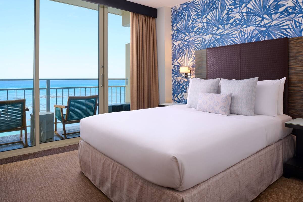 Habitación de hotel con una cama grande, vista al océano, un balcón con sillas y un diseño de papel tapiz tropical.