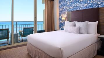 Habitación de hotel con una cama grande, vista al océano, un balcón con sillas y un diseño de papel tapiz tropical.